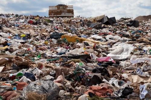 A vast and chaotic landscape filled with a large accumulation of mixed household and commercial waste, including plastic bags, cardboard boxes, paper, broken furniture, and assorted discarded items scattered across an open land area. The debris covers the foreground and midground, creating a dense, unorganized pile of rubbish with various textures and colors, predominantly brown, grey, and black, with occasional brighter tones from plastic and packaging. In the background, a partially constructed or abandoned building structure is visible, with exposed bricks and an incomplete appearance, situated on a slight elevation amid the waste. The scene is set outdoors under a cloudy sky, suggesting daytime with diffused natural lighting. This image illustrates the importance of professional rubbish removal services such as those provided by Garden Clearance Shoreditch, highlighting the scale and mess associated with improper waste disposal in urban environments like Shoreditch, London postcode area.