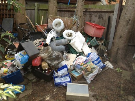 A cluttered garden area with various types of household rubbish and waste materials scattered across the ground, including black and transparent plastic bags filled with debris, white plastic pipes, a large red plastic container, a black plastic crate, and loose packaging. There are also cardboard boxes, plastic bottles, and miscellaneous household items. The waste is piled in front of a damp wooden fence, with several mature trees and a wooden fence visible in the background, suggesting a backyard setting in Shoreditch. The scene appears to be the result of a garden clearance service by Garden Clearance Shoreditch, focusing on rubbish removal and clearing junk from outdoor spaces. The ground is bare soil with some grass patches around the edges, and natural daylight illuminates the area, highlighting the variety of waste materials and the disorganized nature of the rubbish stack.