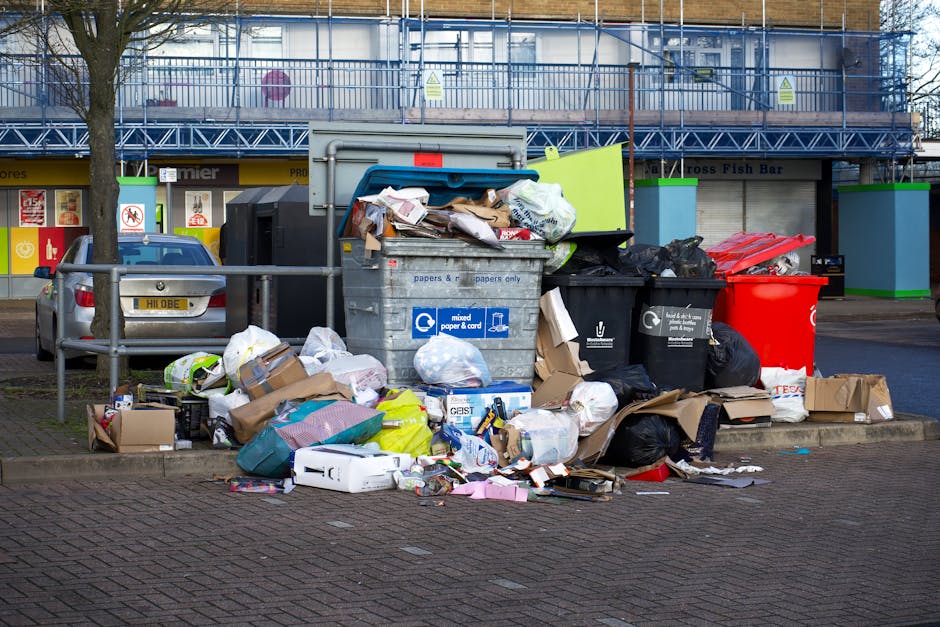 A narrow urban alleyway cluttered with various discarded waste materials, including large black trash bags, cardboard boxes, and scattered debris on the rough concrete ground. The trash bags appear fu