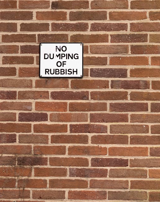 A large, dirty, beige plastic rubbish bag placed on a narrow urban street sidewalk, partially leaning against a weathered, graffiti-marked brick wall with metal bars on the windows. The bag appears to