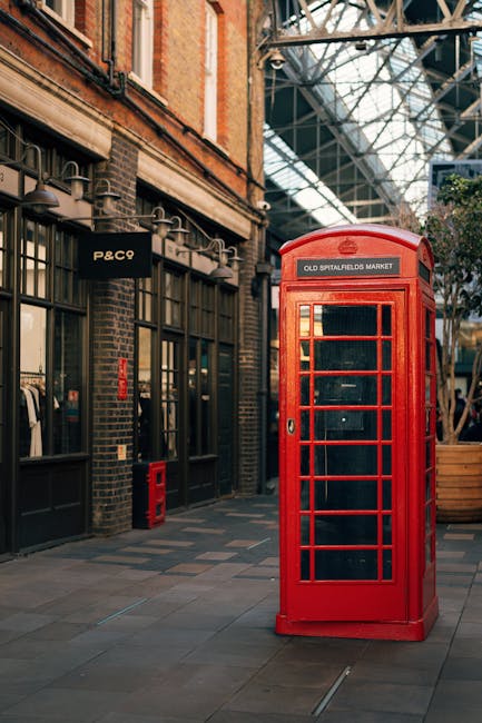 A traditional red British telephone kiosk with a rectangular shape and glass paneled door, positioned on a paved walkway within an indoor market setting. The kiosk features a curved top with a white s